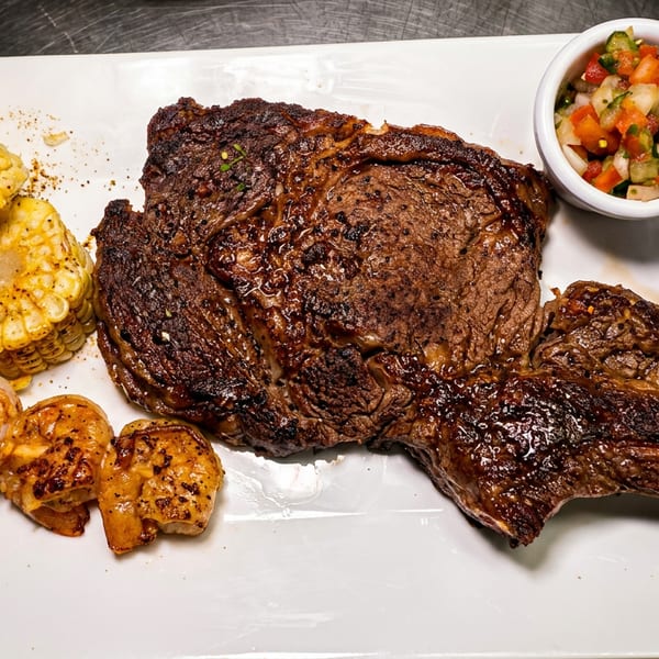 Grilled steak, shrimp skewer, corn on the cob, pico de gallo, and guacamole on a white plate.