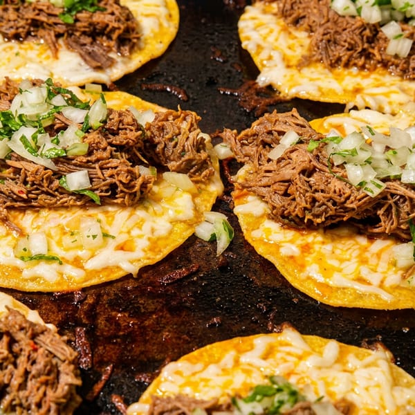 Several birria tacos with shredded beef, melted cheese, and diced onions cooking on a griddle.