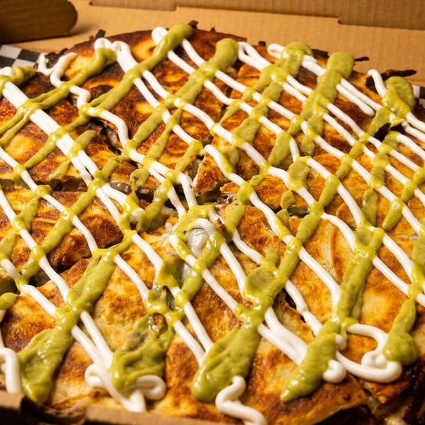 A large quesadilla, cut into wedges, drizzled with green and white sauces in a cardboard box.