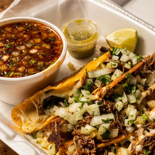 Birria tacos with consommé, green salsa, and a lime wedge in a white takeout container.