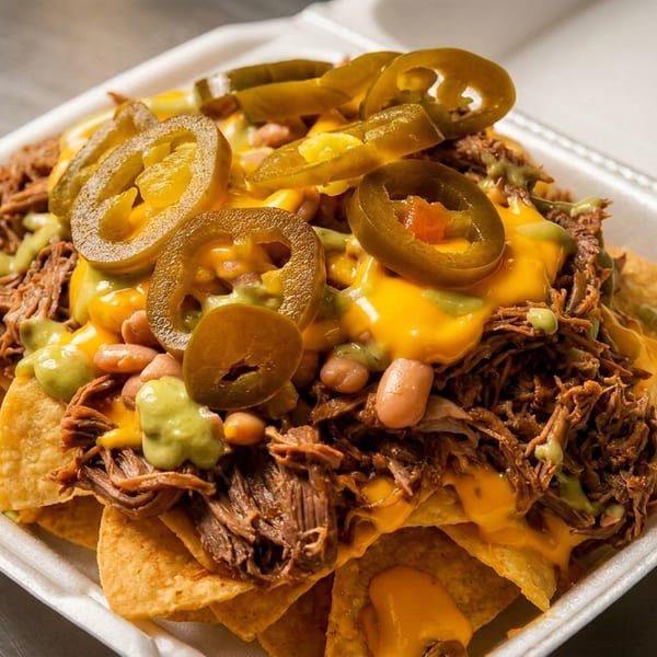 Loaded nachos with shredded meat, cheese sauce, pinto beans, guacamole, and sliced jalapeños in a to-go container.