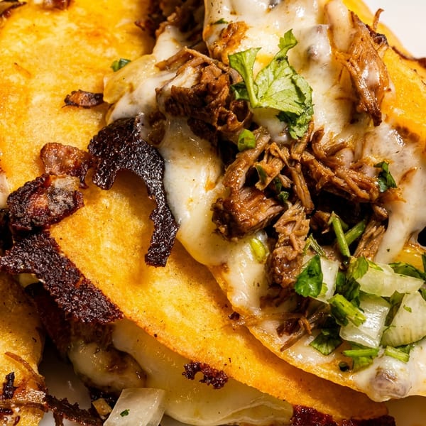 Close-up of three cheesy birria tacos with shredded beef, cilantro, and onions on a white plate.