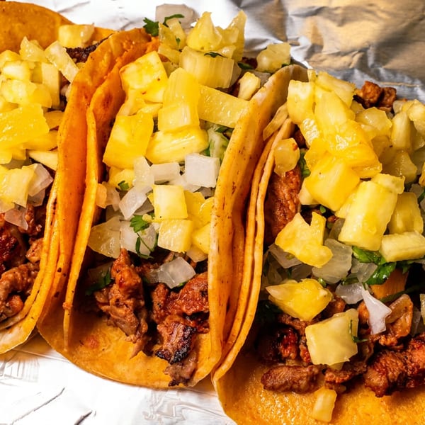 Three al pastor tacos with pineapple and onions on corn tortillas, resting on foil.
